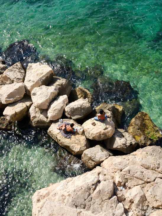 Two people relaxing on rocks overlooking turquoise Mediterranean waters at Catalan beach in Marseille.