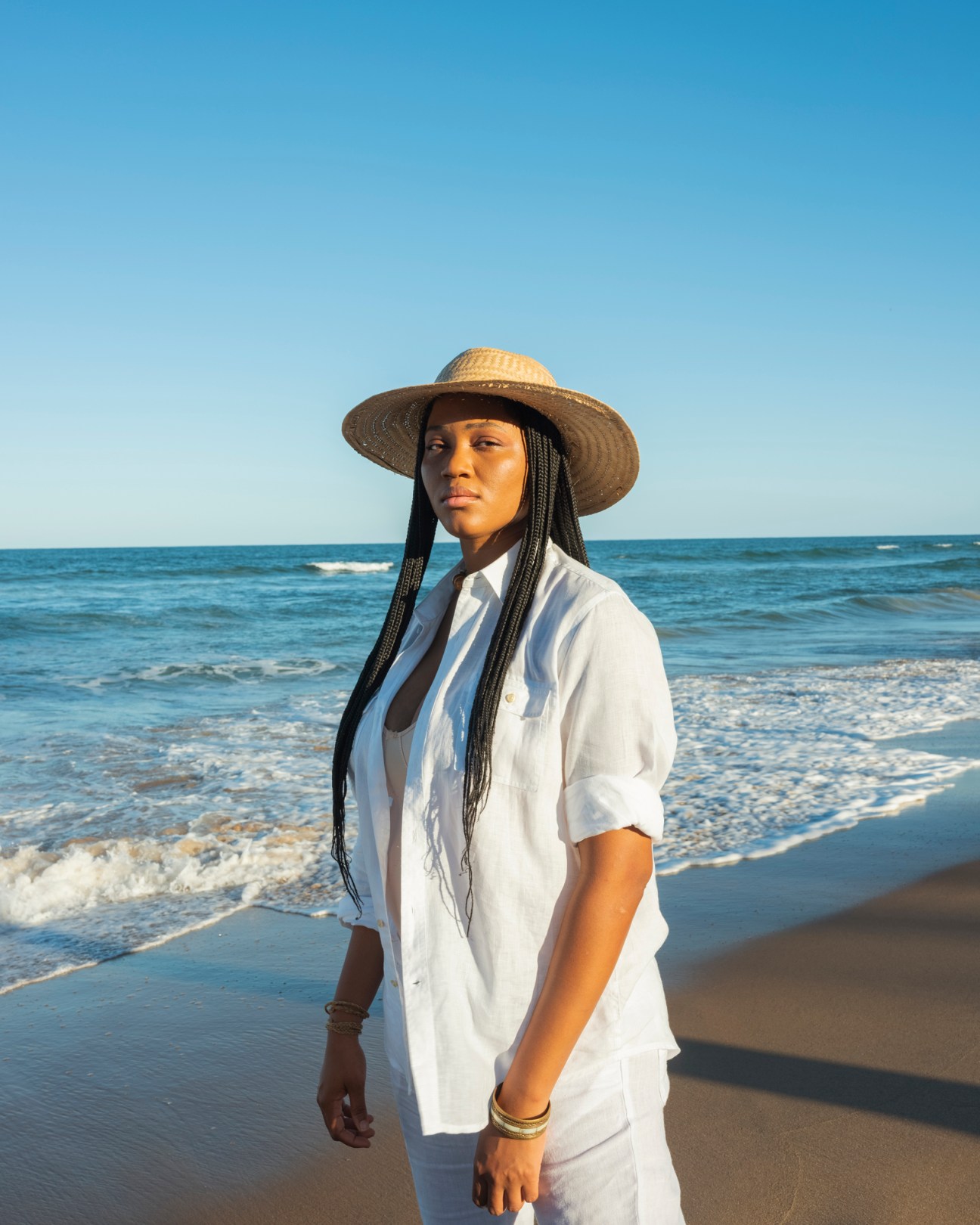 Luedji Luna wearing a white linen shirt and straw hat standing on a beach.