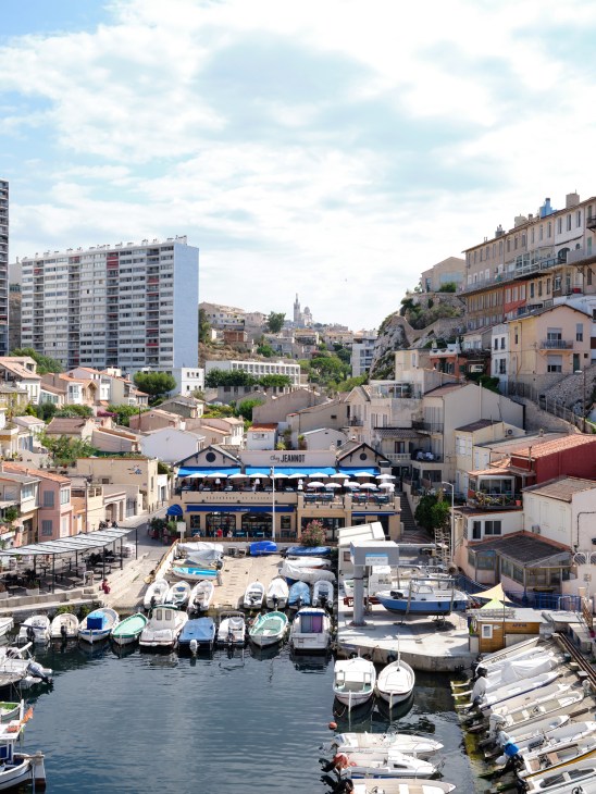 Marseille harbor view with moored boats, waterfront restaurants, and hillside architecture under blue sky.
