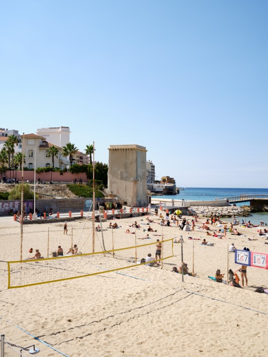 Catalan beach in Marseille with volleyball nets, swimmers, and historic architecture along the shoreline.