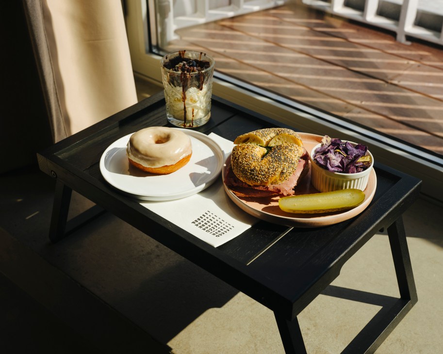 Room service tray with bagel, sandwich, doughnuts, pickles and beverage on black table by window.