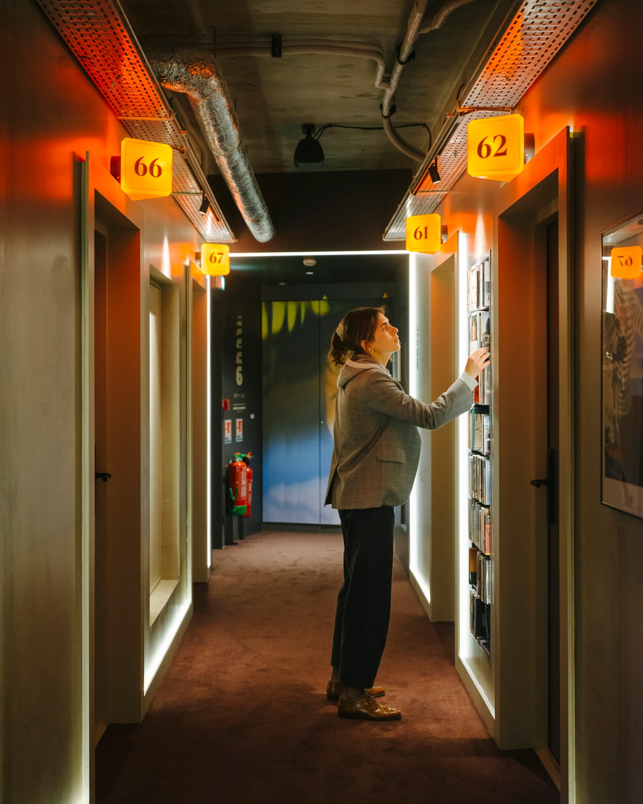 Person browsing self-service DVD shelves lining a hotel corridor with numbered yellow signs and neon lighting.