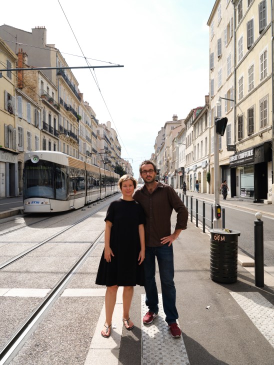Marie-Pierre Gracedieu and Adrien Servières standing on a Marseille street with a tram and historic buildings behind them.