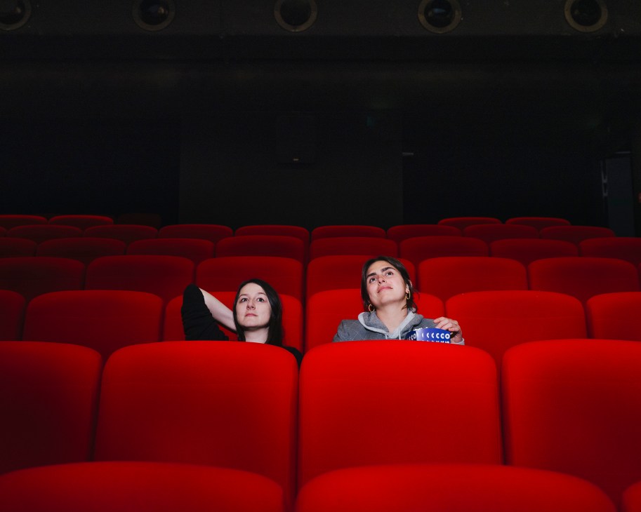 Two women sit among red cinema seats in an empty theatre auditorium.