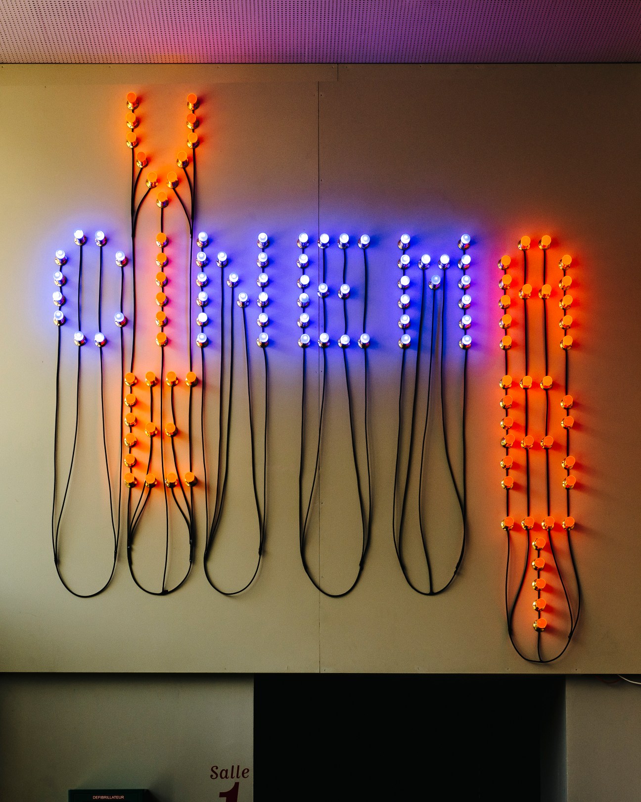 Christian Boltanski neon light installation with white and orange bulbs on black wires against a wall.