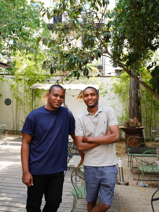 Eric and Hugues Mbenda stand in a sunny courtyard surrounded by greenery and climbing vines.