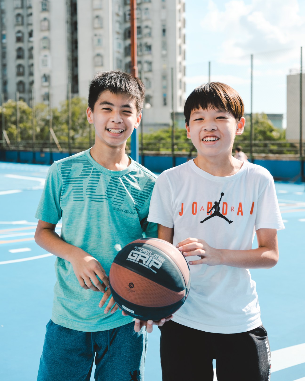 Two smiling boys holding a basketball on a blue outdoor court with apartment buildings in background