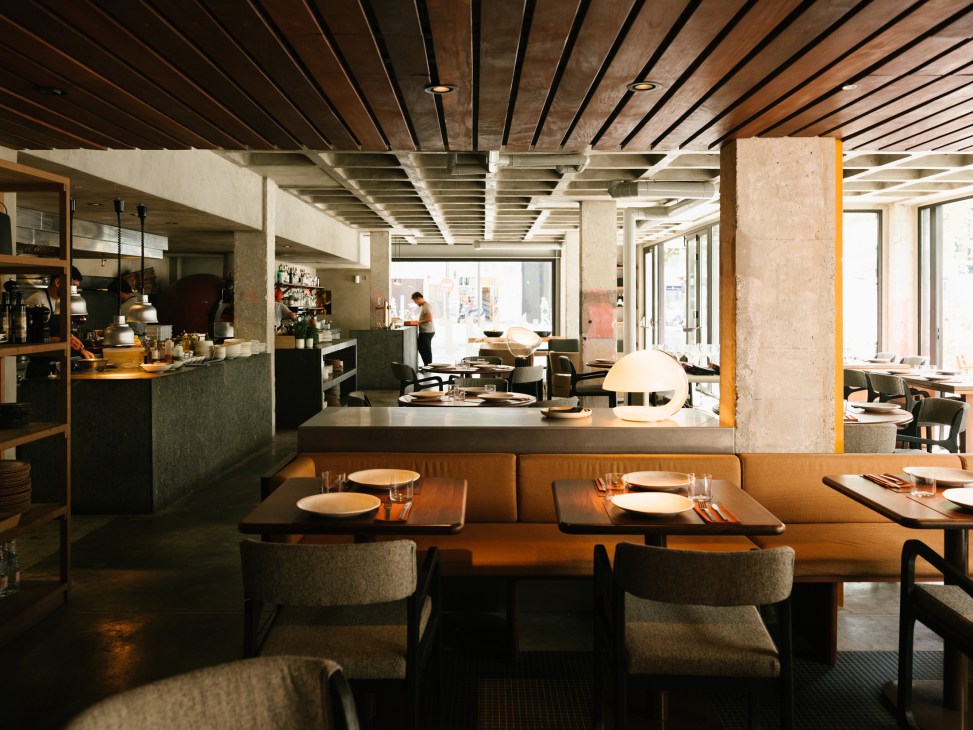 Modern restaurant interior with booth seating along an orange banquette, dark tables, gray upholstered chairs, open kitchen with stone countertop in background, concrete columns, wood-slat ceiling with integrated lighting, and floor-to-ceiling windows overlooking the street.