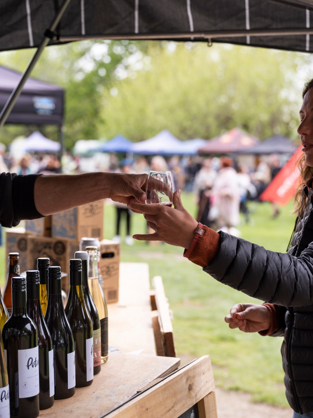 Two people sharing wine glasses at an outdoor farmers market stall with wine bottles displayed on wooden table