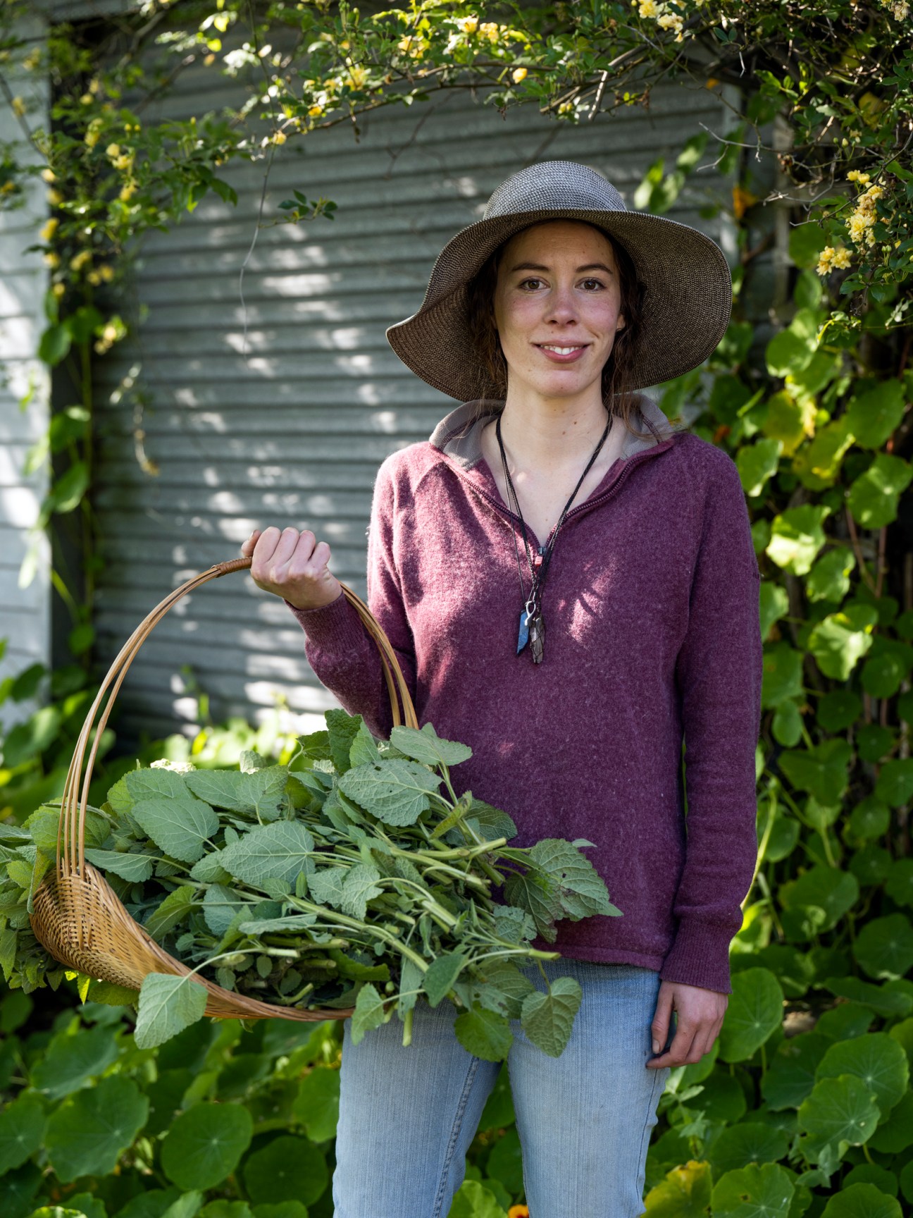 Tea-maker Maya Solomon wearing a wide-brimmed hat and purple sweater, holding a wicker basket full of leafy greens