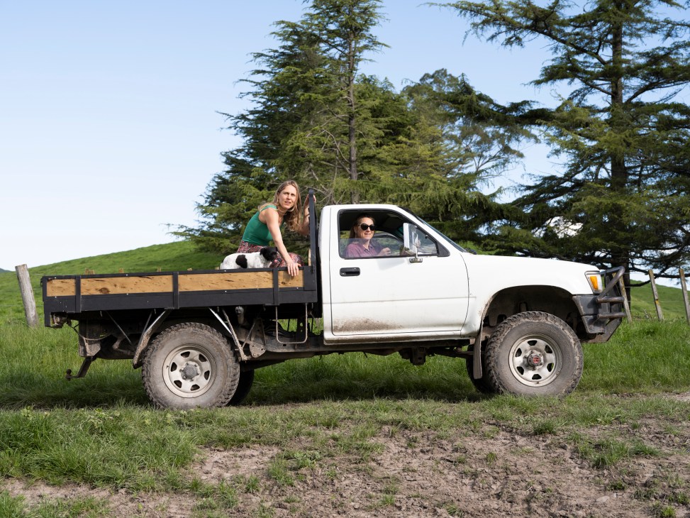 Two women with a white pickup truck on a grassy hillside with trees in the background