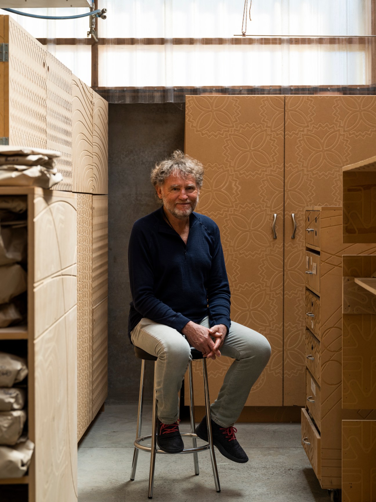 Designer David Trubridge sitting on a stool in his workshop surrounded by wooden furniture and decorative panels