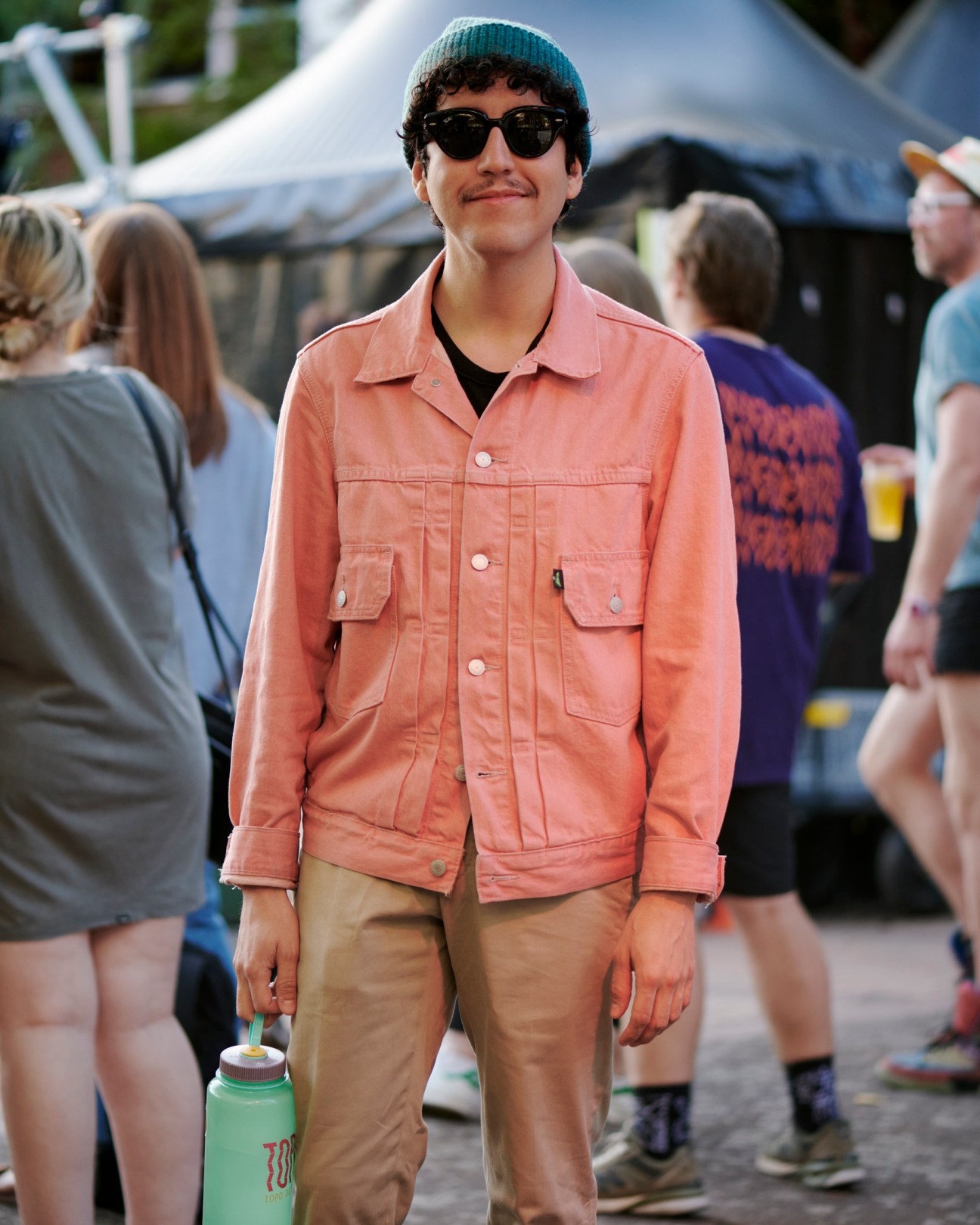 Young man in coral pink jacket, teal beanie and sunglasses at outdoor festival