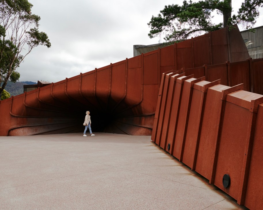 Person walking through a curved rust-colored architectural passageway at Oliver Beer's installation 'Mona Confessional'