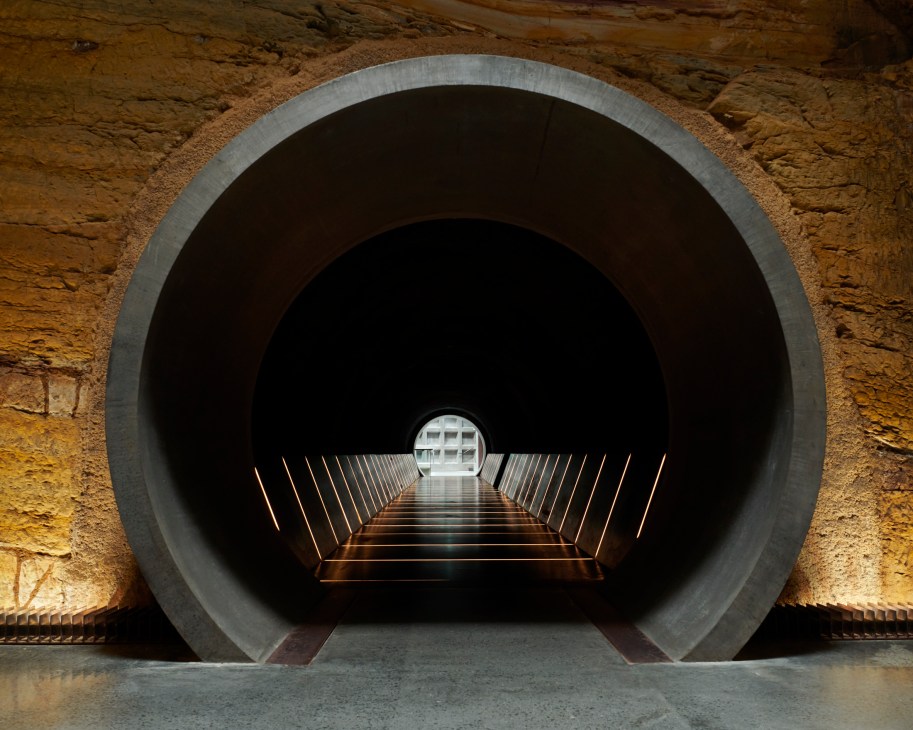 Modern circular tunnel entrance with illuminated stairs leading up through concrete archway in museum setting