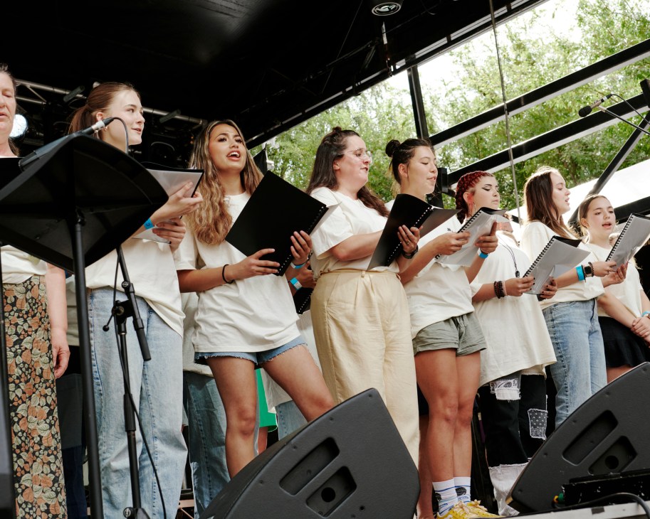 Group of young women in white clothing standing on outdoor stage holding sheet music and singing together