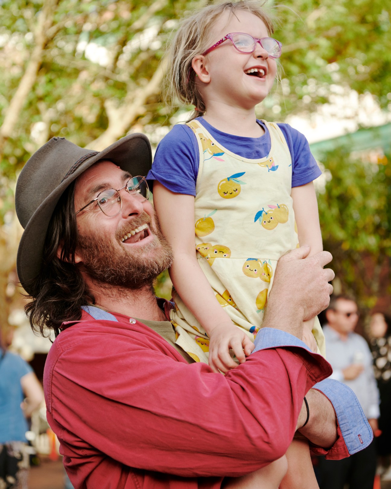 Bearded man in hat and red shirt holding up a young child with glasses at an outdoor festival
