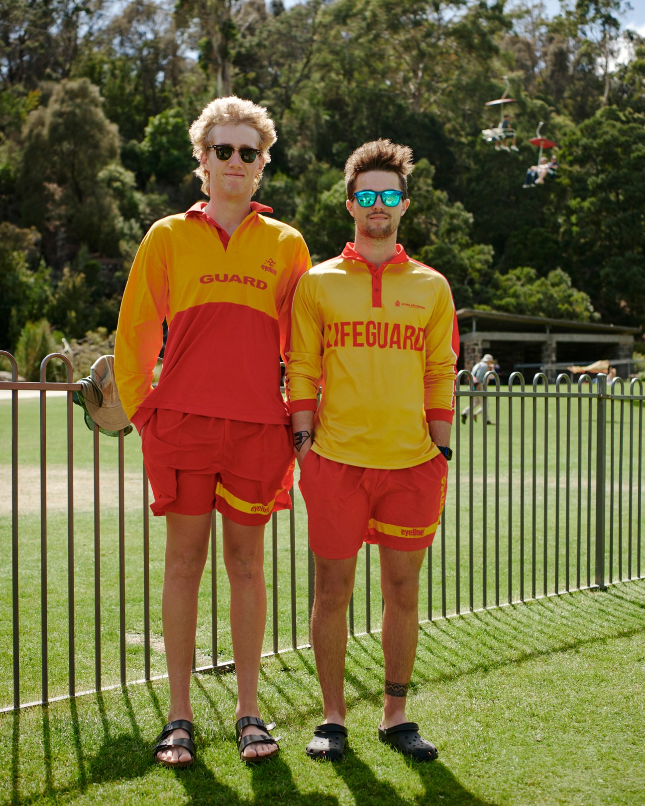 Two people wearing bright yellow and red lifeguard uniforms standing by a fence in a park setting