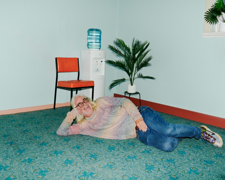 David Walsh lying on patterned carpet in casual clothes next to office equipment and plants
