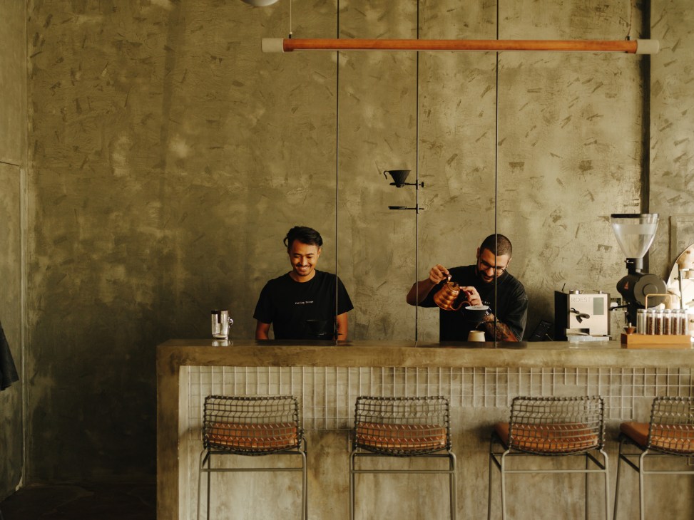 Two baristas prepare coffee at a minimalist counter in a specialty coffee shop with concrete walls.
