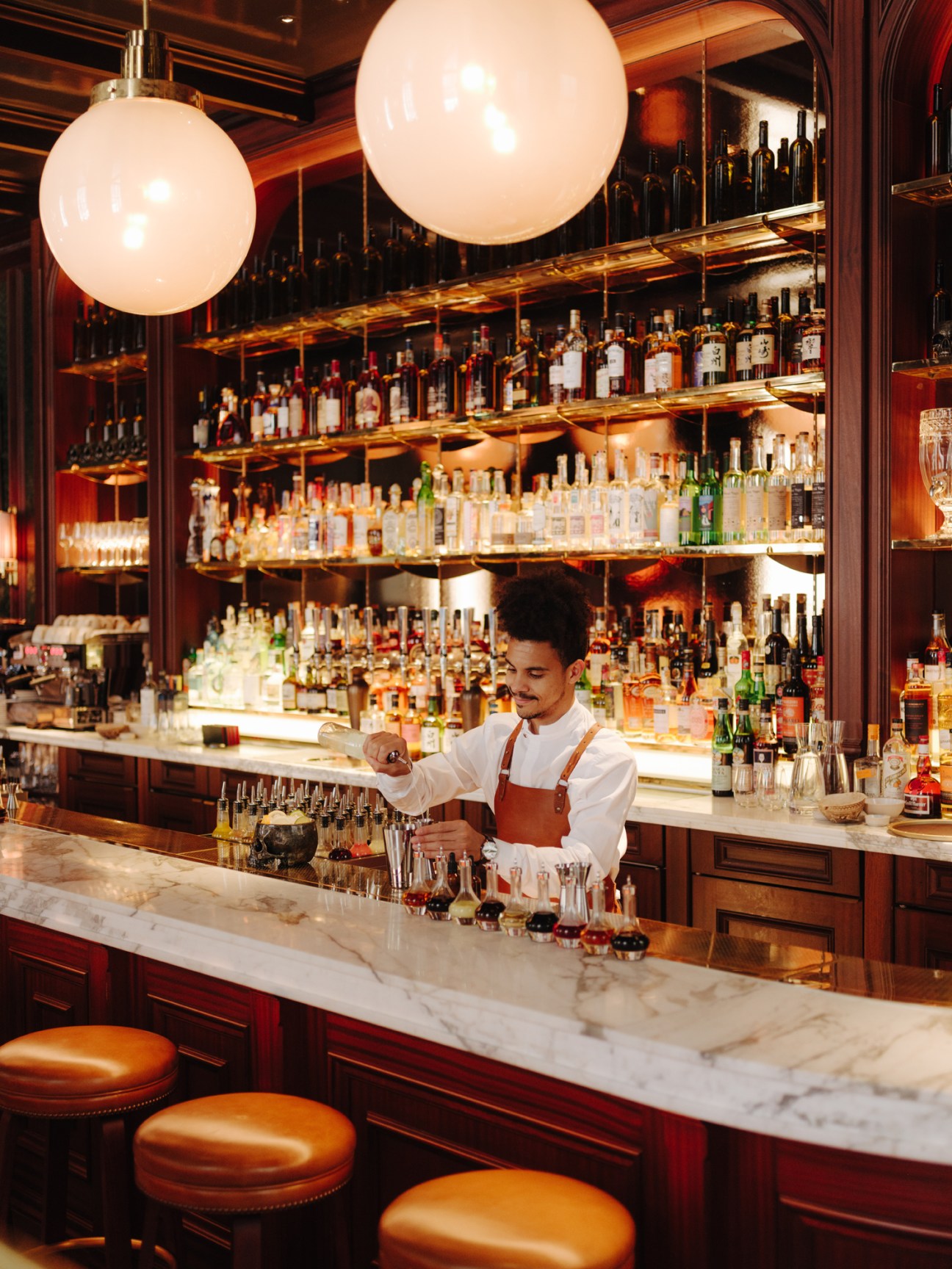 Bartender mixing a cocktail at Side Hustle bar with illuminated shelves of spirits behind him.