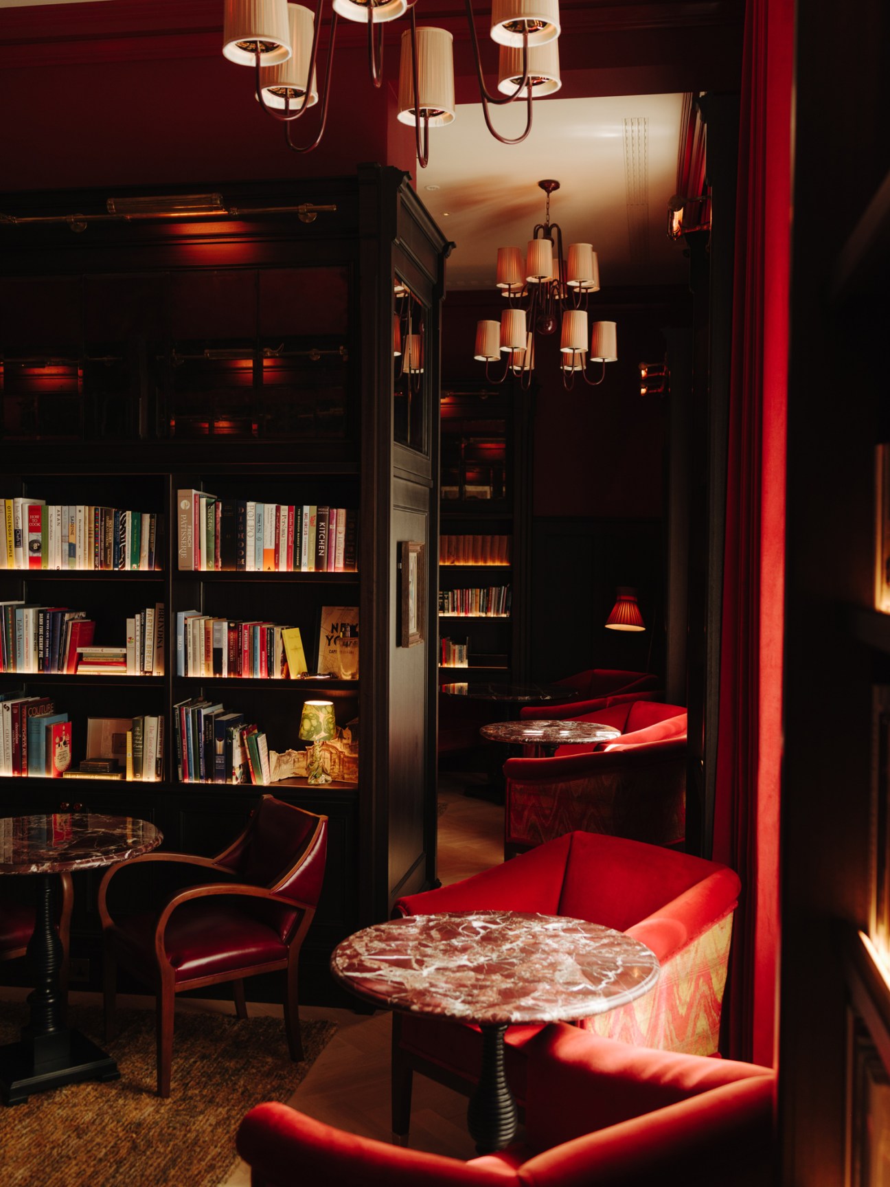 Dimly lit lounge area with red velvet seating, bookshelves, and pendant lighting at Nomad London hotel.