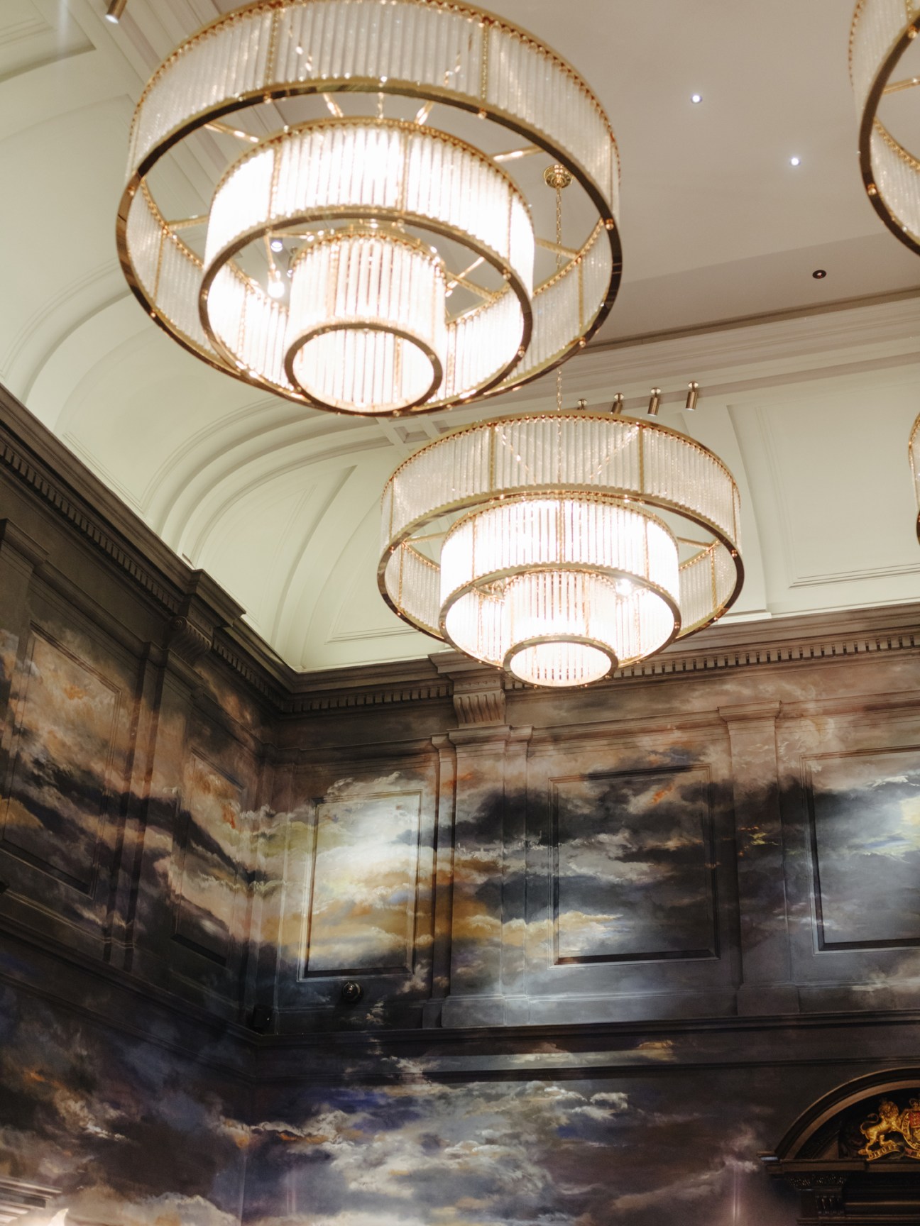 Tiered brass and glass ceiling fixtures illuminating dramatic cloud-themed wall art in the Nomad hotel's interior.