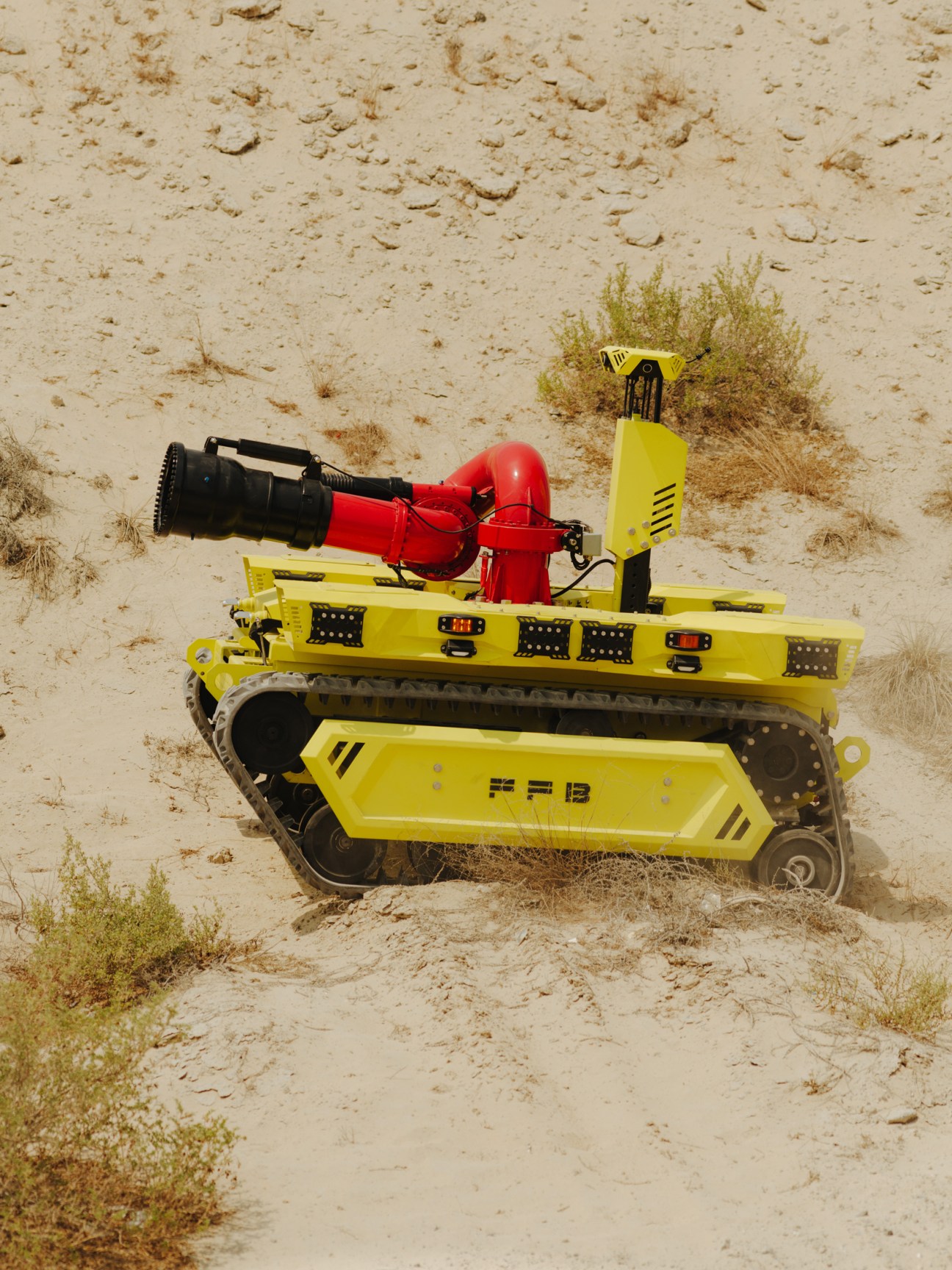 FFBots' yellow tracked firefighting robot with red water cannon on sandy ground with sparse vegetation