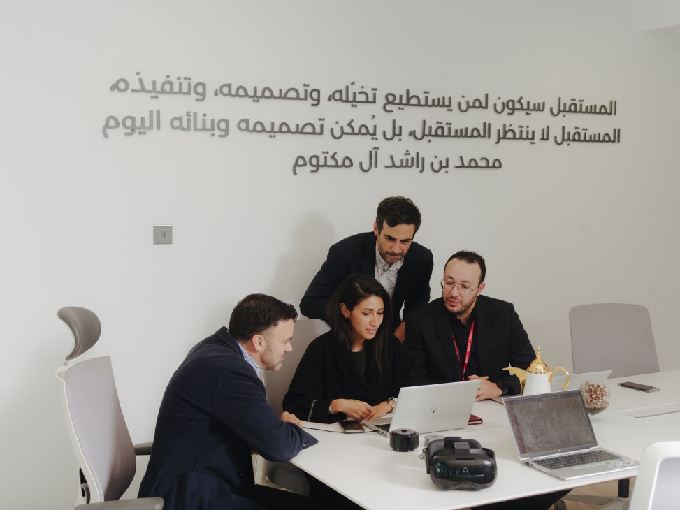 Four professionals in business attire gathered around a white table with laptops and devices, discussing technology in a modern office setting.
