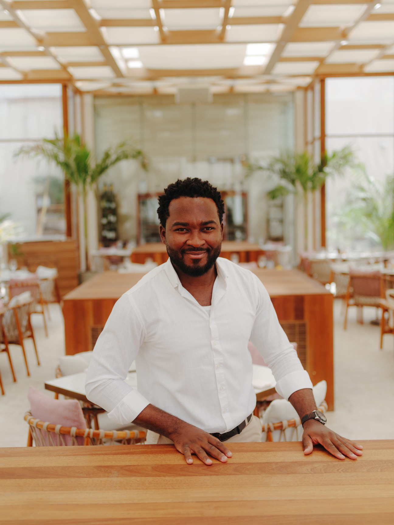 Man in white shirt leaning against wooden counter in modern restaurant with geometric ceiling and palm plants.