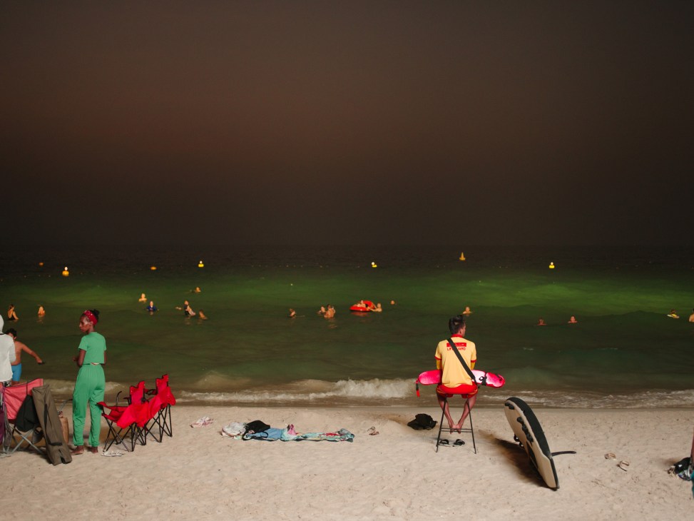 Floodlit Night Swimming Beach in Umm Suqeim with swimmers in illuminated water and beachgoers on sand at dusk.