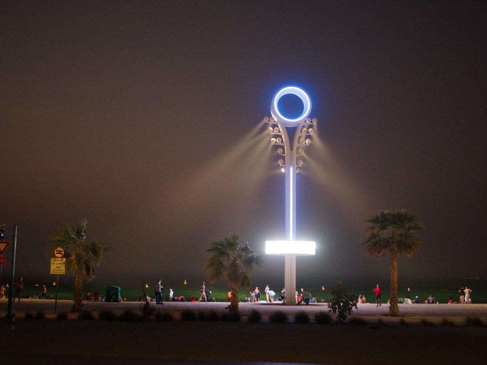 Illuminated blue and white lamp post on Night Swimming Beach at dusk with palm trees and beachgoers.