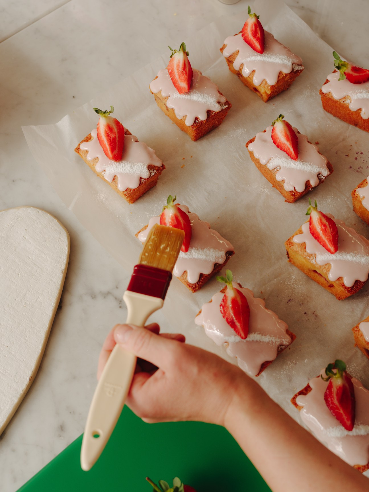 Hand with paintbrush glazing strawberry-topped pastry squares on parchment paper.