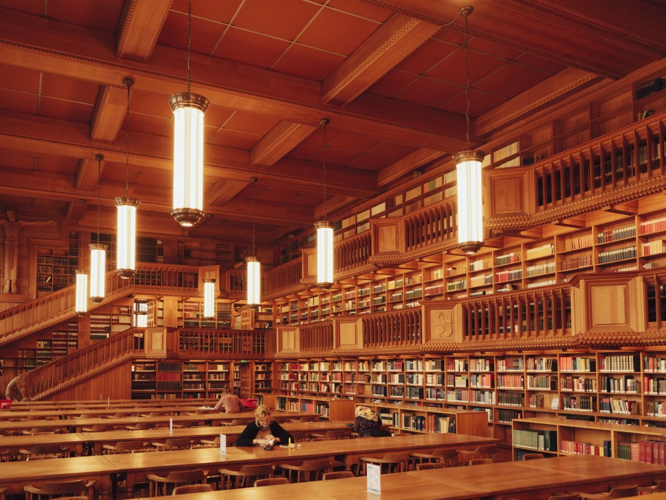 Wooden reading room with red ceiling, hanging cylindrical lights, and multi-level bookshelves filled with books.