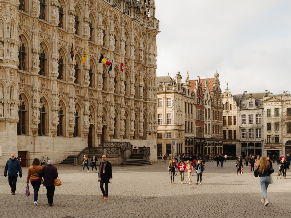 Ornate Gothic town hall with decorative stone facade and Belgian flags in Leuven's Grote Markt square.