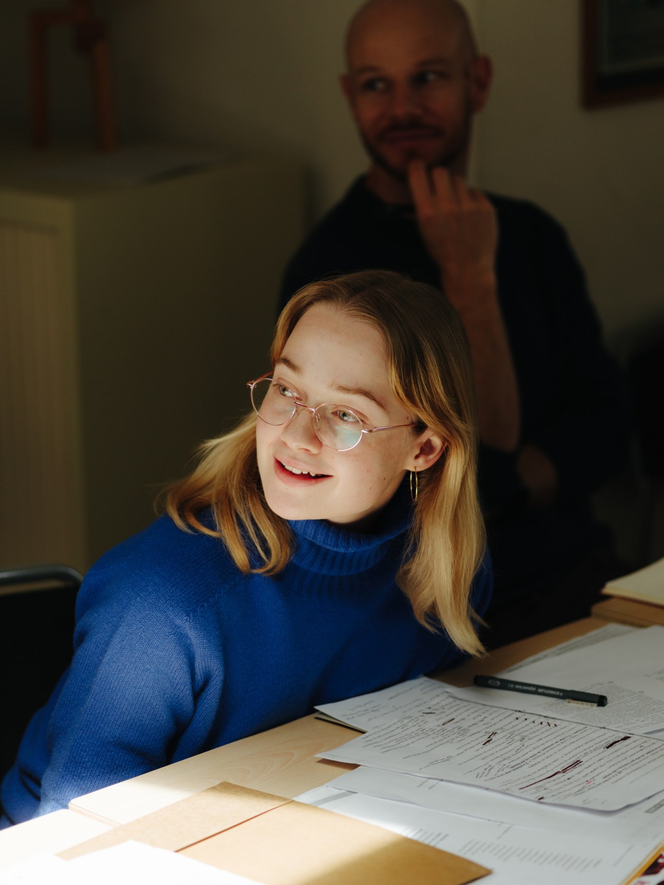 Young woman with glasses and blue sweater studying at desk with papers and books in classroom
