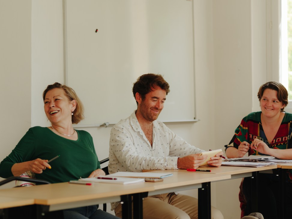 Three language students sitting at desks in a bright classroom, smiling and laughing during a French lesson