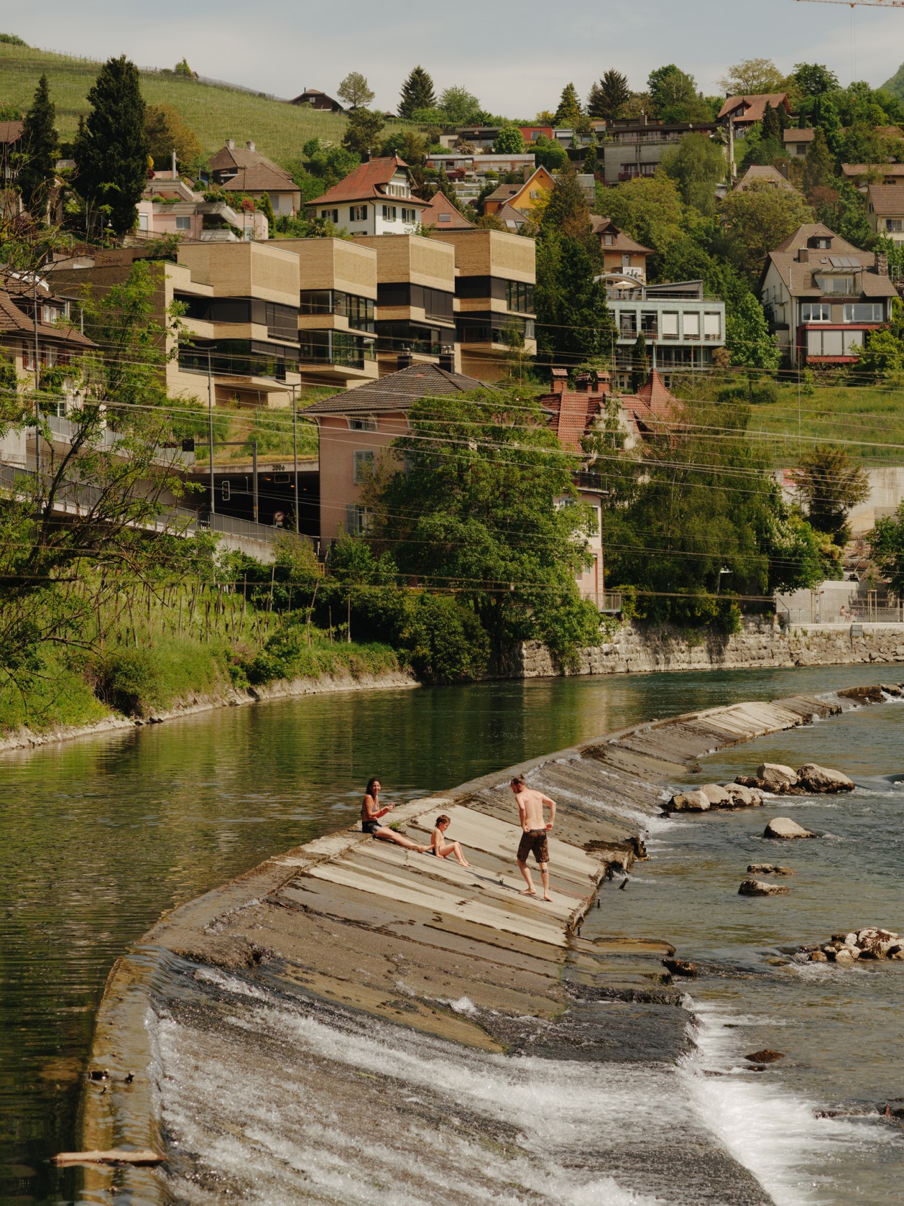 People swimming and relaxing on concrete steps of a river weir with houses visible on the hillside behind