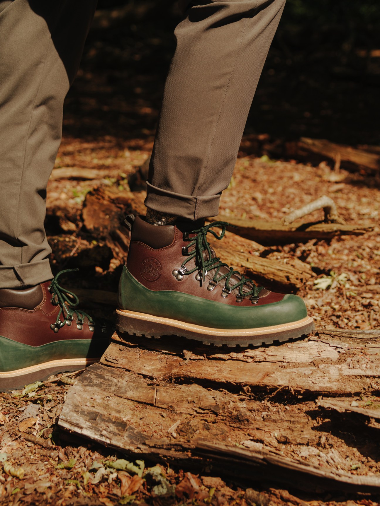 Person wearing brown and green hiking boots walking on fallen logs in autumn forest terrain