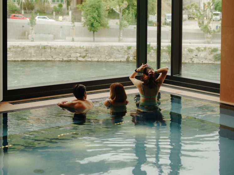Three people relaxing in an indoor thermal pool overlooking the Limmat river through large windows