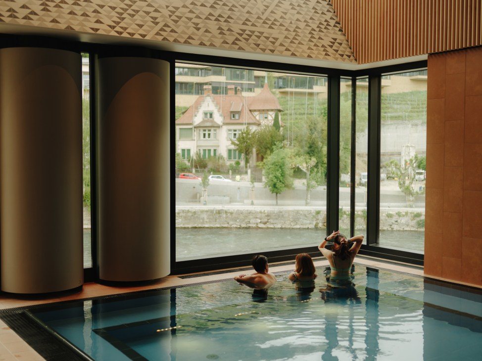 Three people relaxing in an indoor thermal pool overlooking the Limmat river through large windows