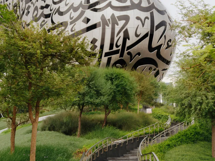 A large spherical white structure with Arabic calligraphy carved into its surface, surrounded by green landscaping and an ascending staircase.