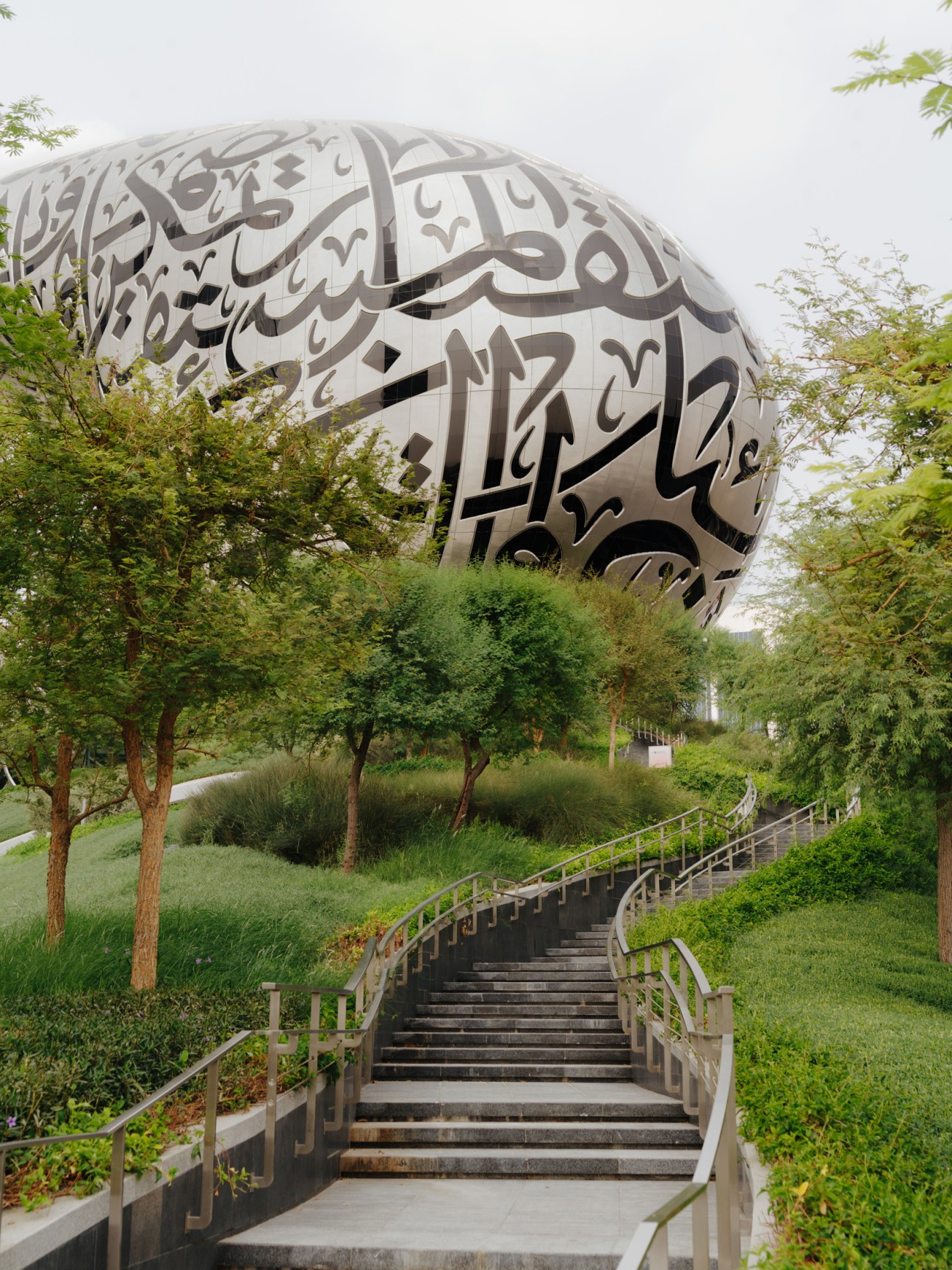 A large spherical white structure with Arabic calligraphy carved into its surface, surrounded by green landscaping and an ascending staircase.