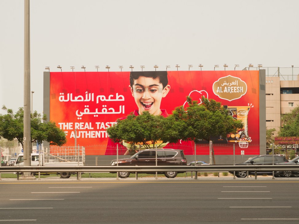 Large red billboard advertising Al Areesh food brand with a smiling boy, partially obscured by a green tree.