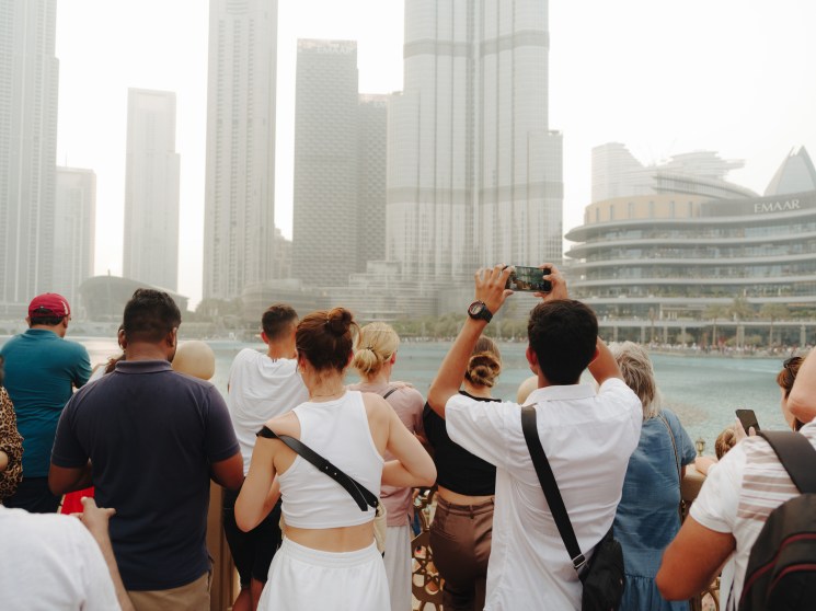 Crowds of tourists photograph Downtown Dubai's skyline and the Burj Khalifa from a waterfront viewpoint.