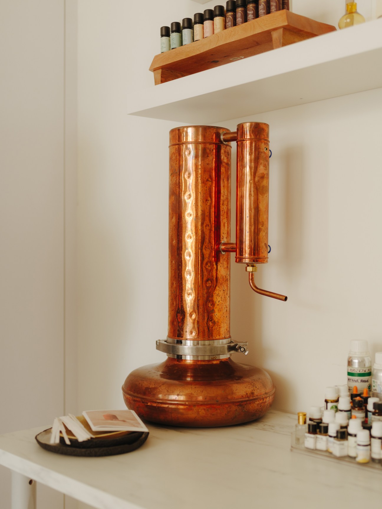 A copper still with distillation apparatus used to extract essential oils and hydrosols, displayed on a white workspace with fragrance bottles on shelves above.
