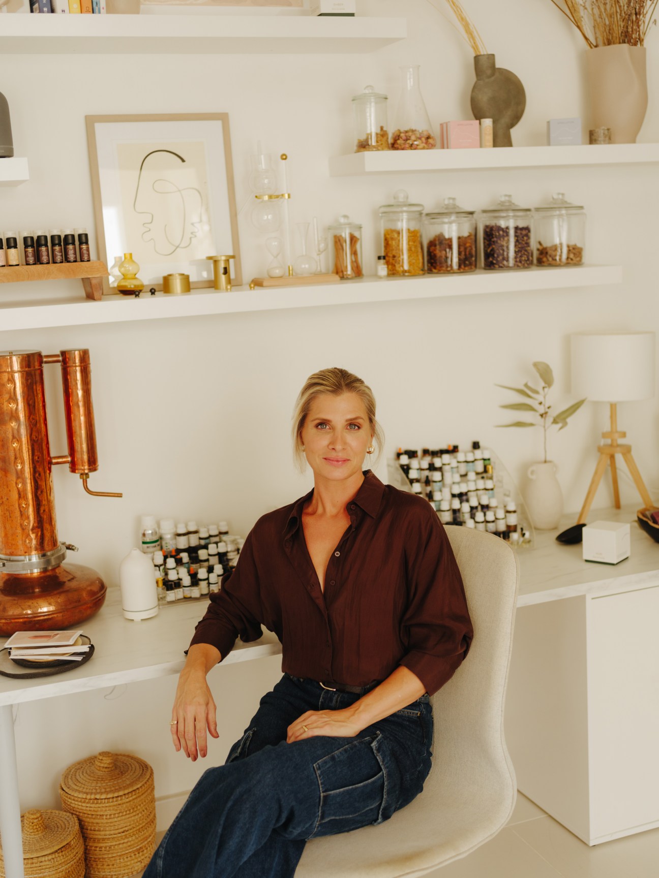 Woman in burgundy shirt and jeans seated at desk in white studio workspace with shelves of fragrance bottles and ingredients.