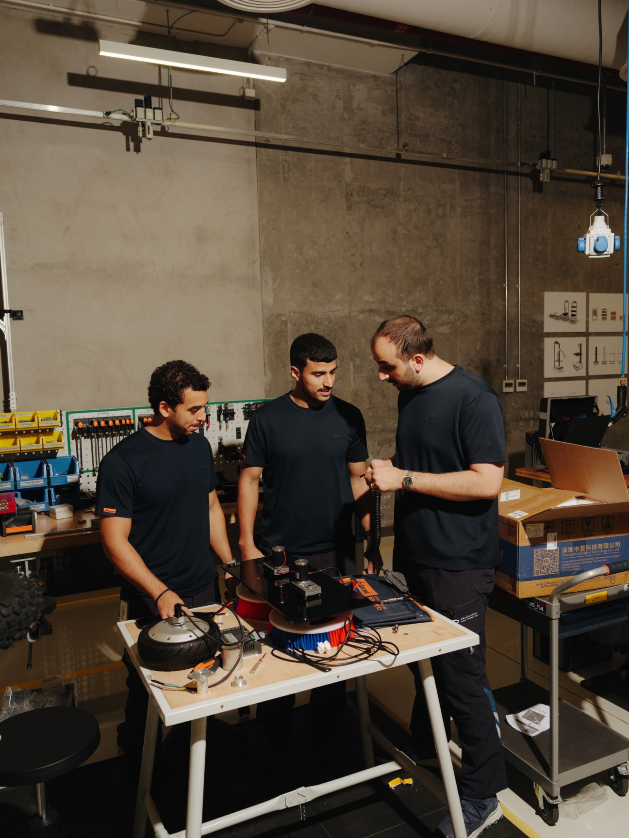 Three engineers in black shirts discuss a robotic project at a workbench in a laboratory space.