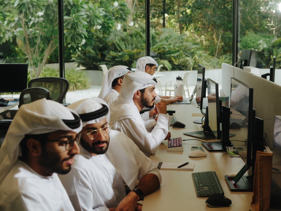 Emirati men in traditional white dress work at computer stations in a modern office with garden views.
