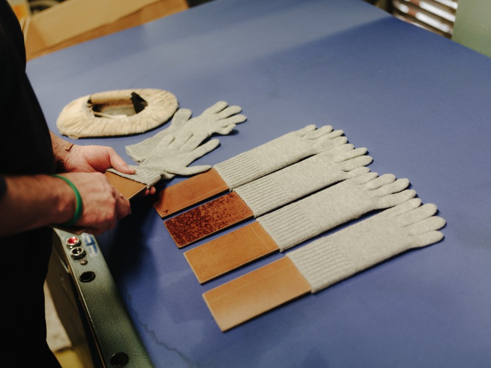 Worker examines cream-colored cashmere gloves and fabric swatches on a blue table.